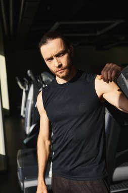 Aktif giyinmiş atletik bir adam spor salonundaki bir dizi koşu bandının yanında duruyor, antrenman rutinine hazır..