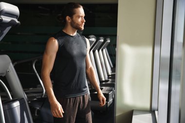Aktif kıyafetli bir adam spor salonundaki bir sıra koşu bandının önünde özgüvenle duruyor. Sıkı bir antrenmana hazır..
