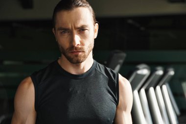 Siyah tişörtlü atletik bir adam spor salonundaki koşu bandının önünde güvenle durur..