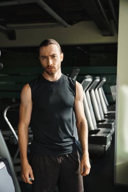 Aktif giyinmiş atletik bir adam bir sıra koşu bandının önünde duruyor, zorlu bir antrenman için hazır..