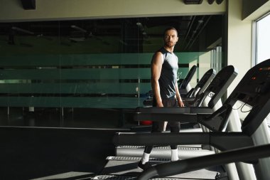 Aktif giyinmiş atletik bir adam spor salonundaki koşu bandında kendine güvenerek duruyor..