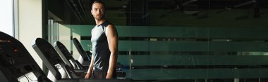 Aktif giyinmiş atletik bir adam bir sıra koşu değirmeninin önünde durur ve antrenman yapmaya hazırdır..