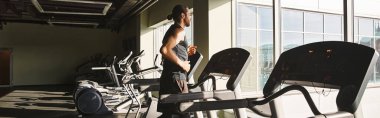 A fit man in athletic wear stands next to a row of exercise bikes in a gym. clipart