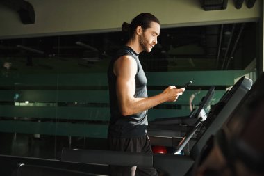 Atletik bir adam koşu bandında ayakta duruyor, spor salonunda spor yaparken cep telefonuyla meşgul..