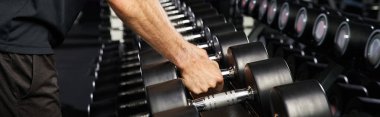 Spor kıyafetli bir atletik bir adam bir çift dambıl tutuyor. Gücünü ve forma bağlılığını gösteriyor..
