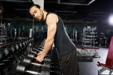 Aktif giyimli formalı bir adam spor salonunda bir sıra dambılın yanında duruyor ve yoğun bir antrenmana hazırlanıyor..