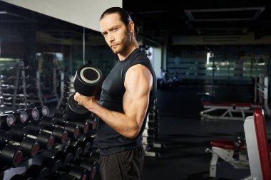 Aktif giyinen atletik bir adam spor salonunda bir dambılı kaldırır, güç ve kararlılık gösterir..