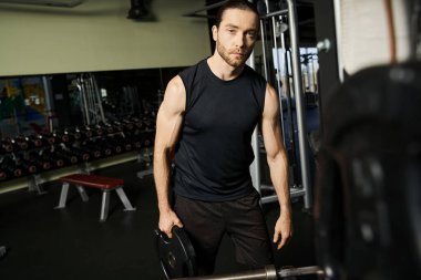 Aktif kıyafet giymiş kaslı bir adam spor salonunda elinde siyah bir tabakla dikiliyor..
