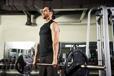Aktif giyinmiş atletik bir adam spor salonunda elinde bir halter, kararlı ve odaklanmış bir şekilde duruyor..