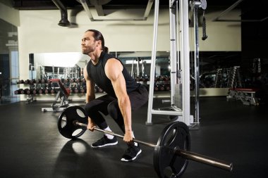 Aktif giyimde odaklanmış bir adam spor salonunda halterle bir çömelme hareketi yapar. Güç ve kararlılık gösterir..
