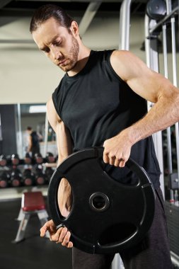 Spor kıyafetleri içinde kaslı bir adam spor salonunda siyah bir tabak tutuyor. Güç ve kararlılık gösteriyor..
