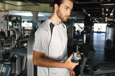 Aktif giyinen atletik bir adam spor salonunda elinde bir şişe su tutarak mola verir..