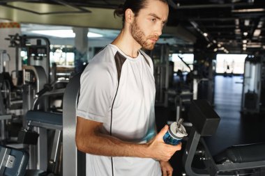 An athletic man in active wear takes a break, holding a bottle of water in a gym surrounded by exercise equipment. clipart