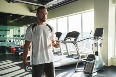 Aktif giyinen atletik bir adam spor salonunda kendine güvenen bir su şişesi tutuyor..