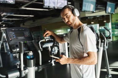 Aktif giyinmiş atletik bir adam koşu bandında koşuyor, kulaklıkları takılı olarak antrenmana dalmış..