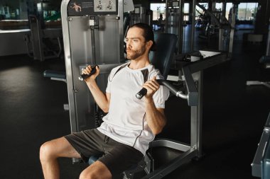 Aktif giyinen atletik bir adam spor salonundaki bir bankta oturur, setler arasında durup odaklanmış bir ifadeyle durur..
