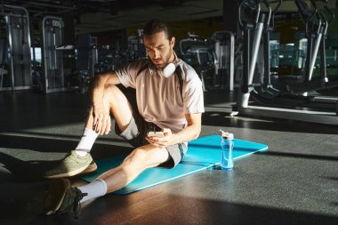 Aktif giyinmiş atletik bir adam spor salonunda mavi bir paspasın üzerinde oturuyor..