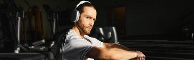 Aktif giyinen odaklanmış bir adam spor salonunda oturup kulaklıkla müzik dinliyor..