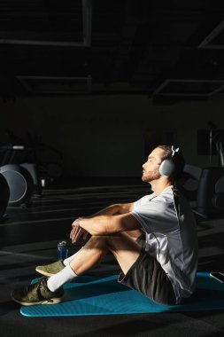 Aktif giyinmiş atletik bir adam bir mindere oturur, kulaklık takar, kendini müziğe veya meditasyona daldırır..