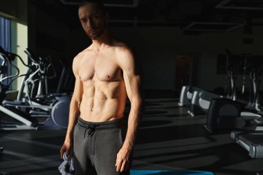 Tişörtsüz kaslı bir adam, modern karanlık bir spor salonunda güvenle duruyor..