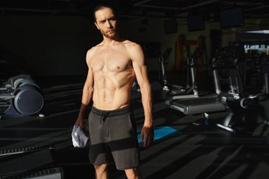 Tişörtsüz kaslı bir adam, spor salonunda kendinden emin duruyor..
