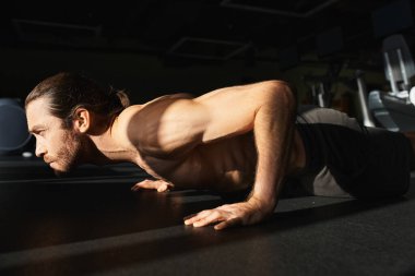 Gömleği olmayan kaslı bir adam spor salonundaki paspasın üzerinde şınav çekiyor, odaklanmış ve kararlı..