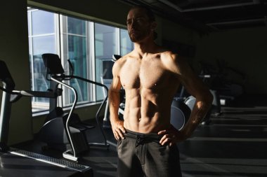 Tişörtsüz kaslı bir adam spor makinesinin önünde duruyor, odaklanmış ve çalışmaya hazır..