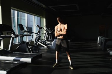 Tişörtsüz kaslı bir adam spor salonundaki koşu bandının önünde güvenle duruyor..
