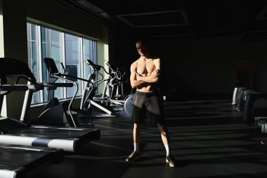 A muscular man without a shirt standing confidently in front of a row of treadmills in a gym. clipart