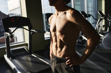 Üstsüz, kaslı bir adam spor salonundaki koşu bandının yanında duruyor..