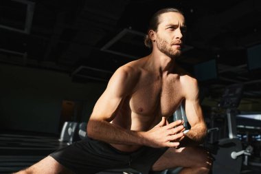 Çıplak göğüslü kaslı bir adam, spor salonundaki bir bankta çömeliyor, forma olan bağlılığını ve gücünü gösteriyor..