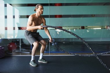 Üstsüz, kaslı bir adam spor salonunda kendine güvenen bir ip tutuyor, gücünü ve kararlılığını gösteriyor..