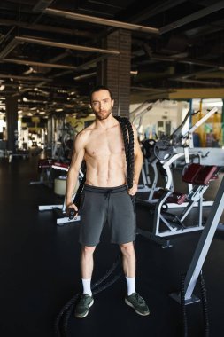 Tişörtsüz kaslı bir adam, spor salonunda dikilmiş, forma ve dayanıklılık eğitimine olan bağlılığını gösteriyor..