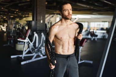 Tişörtsüz, kaslı bir adam ağır iplerle spor salonuna odaklanmış..