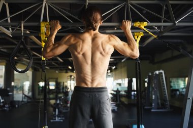 Spor salonunda üstsüz bir adam barfiks çekiyor, kaslı vücudunu ve forma bağlılığını gösteriyor..