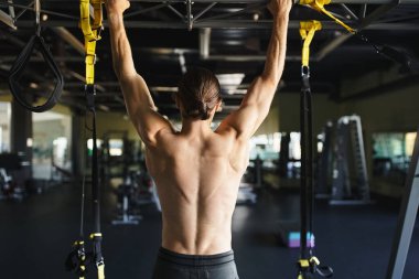 Üstsüz, kaslı bir adam egzersiz malzemeleriyle dolu bir spor salonunda barfiks çekiyor..