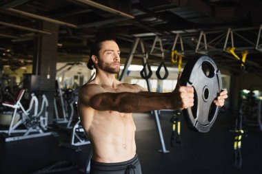 Üstsüz bir adam spor salonunda halter kaldırır kaslı fiziğini ve forma bağlılığını gösterir..