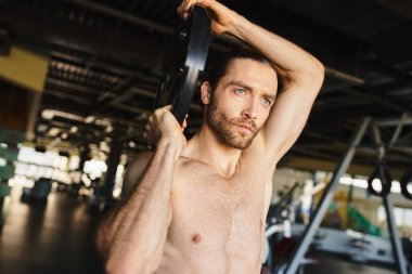 Tişörtsüz bir adam spor salonunda ağırlık levhasını kavrayıp kaslı vücudunu gösterir ve egzersiz rutinine odaklanır..