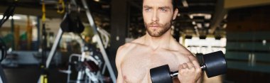 Spor salonunda dambılı tutarken kaslı vücudunu gösteren üstsüz bir adam..