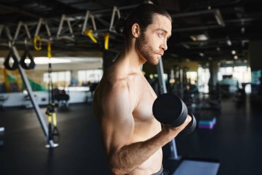 Spor salonunda elinde dambıl tutan, kaslı fiziğini ve forma olan bağlılığını gösteren üstsüz bir adam..