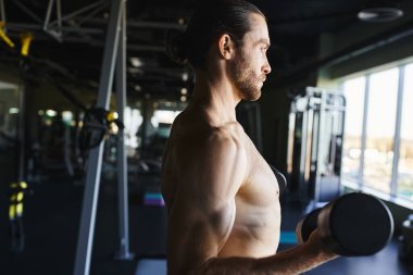 Spor salonunda bir dambılı tutarken kaslarını esneten üstsüz bir adam antrenman rutinine bağlılığını gösteriyor..
