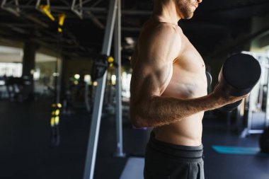 Gömleksiz bir adam spor salonunda dambılı tutarken kas tanımı sergiliyor..