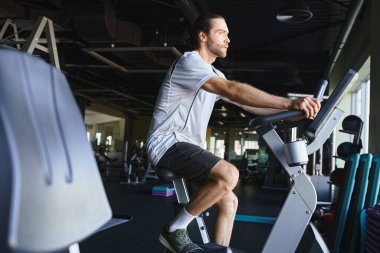 Kaslı bir adam spor salonunda durgun bir bisikletin üzerinde enerjiyle bisiklet sürüyor..