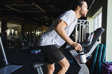 Kaslı bir adam spor salonunda sabit bir bisiklete biniyor, ham güç ve kararlılık sergiliyor..
