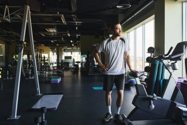 Spor salonunda duran kaslı bir adam. Etrafı egzersiz malzemeleriyle çevrili..