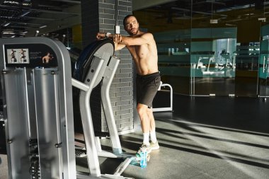 Tişörtsüz kaslı bir adam spor salonunda spor makinesinin yanında duruyor ve egzersiz yapmaya hazırlanıyor..