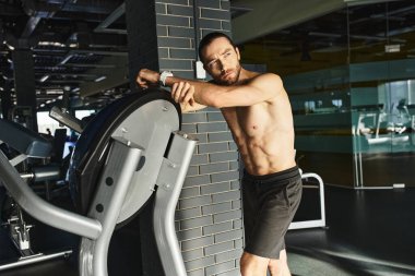 Üstsüz, kaslı bir adam spor salonundaki spor makinesinin yanındaki duvara yaslanıyor..