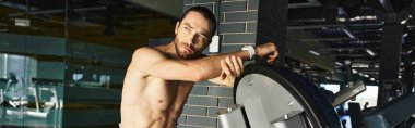 Tişörtsüz, kaslı bir adam spor salonunun duvarına yaslanmış, heykel şeklini sergiliyor..
