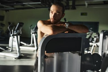 Spor salonunda spor yaparken makineye yaslanan üstsüz, kaslı bir adam..