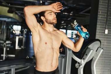 Güçlü üstsüz adam mola veriyor, spor salonunda elinde bir şişe su tutuyor..
